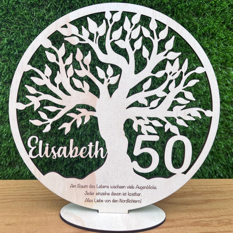 May include: A wooden, circular tree of life decoration with the name "Elisabeth" and the number "50" cut out. The tree has many leaves and is mounted on a round base. Text at the bottom reads in German.