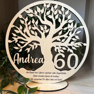 May include: A wooden, circular decorative piece featuring a tree design with the name "Andrea" and the number "60" cut out. The piece includes a base for standing. Text in German is also present.