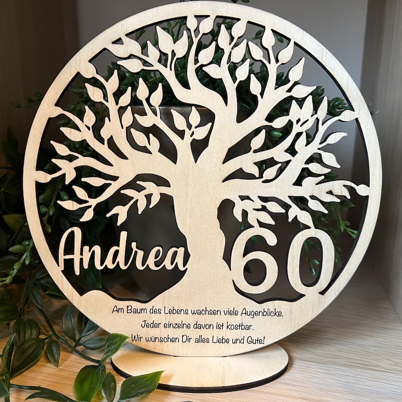 May include: A wooden, circular decorative piece featuring a tree design with the name "Andrea" and the number "60" cut out. The piece includes a base for standing. Text in German is also present.