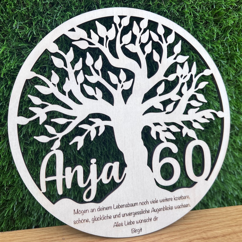May include: A round, wooden tree of life plaque with the name "Anja" and the number "60" cut out. The plaque has a natural wood finish and includes a message and the name "Birgit" at the bottom.