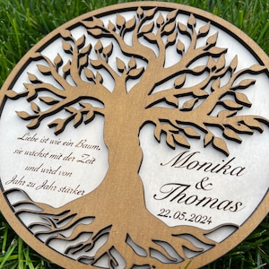 Könnte beinhalten: Runde Holzplakette mit einem Baum des Lebens, hellbraune Oberfläche. Das Design zeigt einen Baum mit aufwendig ausgeschnittenen Blättern und Wurzeln. Die Namen "Monika & Thomas" und das Datum "22.05.2024" sind eingraviert. Der Durchmesser beträgt ca. 20 cm.
