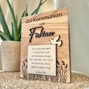 Könnte beinhalten: Holztafel mit dem Text "Zur Kommunion von Fabian" und einem ausgeschnittenen Taubenmotiv. Die Tafel enthält eine gedruckte Botschaft und das Datum "03.Mai 2026". Das Design zeigt graviertes Gras und eine natürliche Holzmaserung.