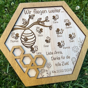 Könnte beinhalten: Holztafel in sechseckiger Form mit hellbraunem Rahmen. Die Tafel zeigt ein Bienen-Design mit einem Bienenstock, Bienen und dem Text "Wir fliegen weiter..." und Namen. Zusätzlicher Text beinhaltet "Liebe Anna, Danke für die tolle Zeit!" und "Kita 2022/2023".