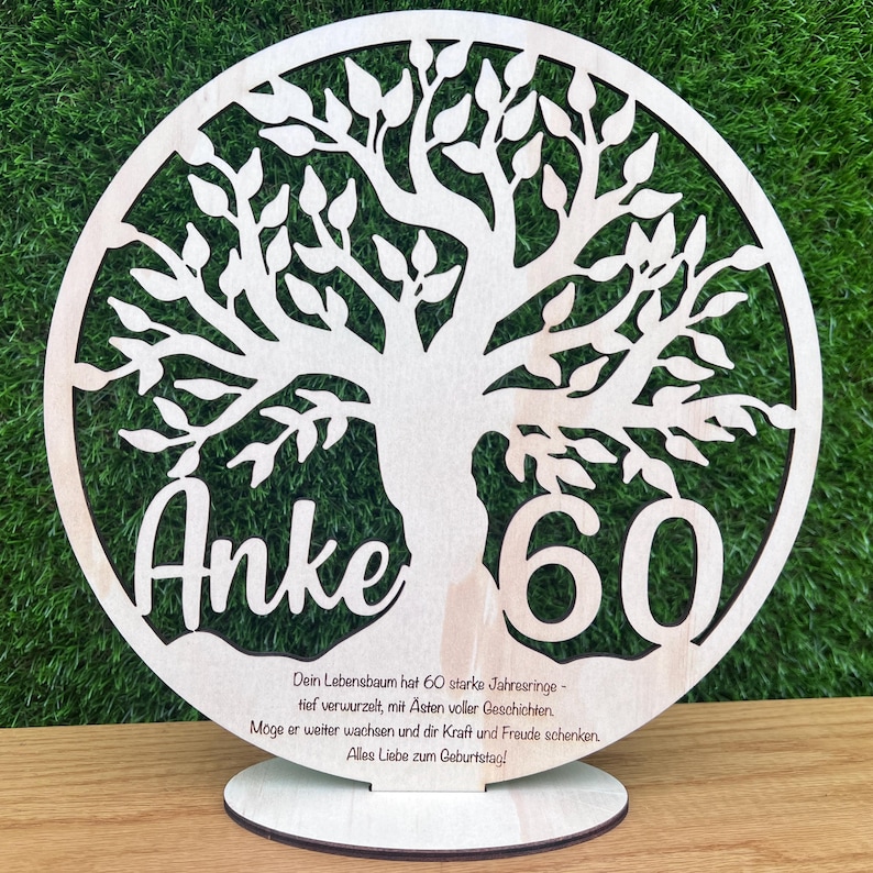 May include: A wooden tree-shaped decoration with the name "Anke" and the number "60" cut out. The tree is inside a circular frame and has a base for standing. The text below reads in German.