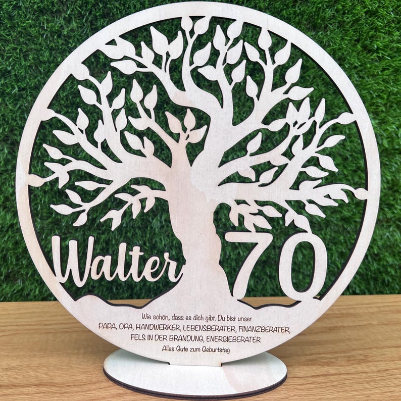 May include: A wooden, laser-cut tree of life design with the name "Walter" and the number "70". The base includes text in German. The tree is set against a green background.