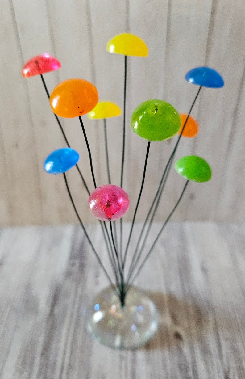 K&ouml;nnte beinhalten: Eine bunte Glasblumen-Arrangement mit neun runden, durchsichtigen Blumen in verschiedenen Farben, darunter rosa, orange, gelb, gr&uuml;n und blau. Die Blumen sind an d&uuml;nnen schwarzen Dr&auml;hten befestigt und in einer klaren Glasvase angeordnet.