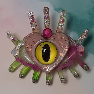 May include: A whimsical, handmade brooch featuring a central yellow eye with a black pupil, surrounded by a heart-shaped design in pink and glitter. The brooch has multiple crystal and glitter accents, with green and purple elements.