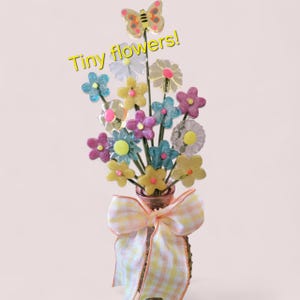 Könnte beinhalten: Ein Strauß bunter, winziger Blumen in einer kleinen Vase, gebunden mit einem gelb-weiß karierten Band. Die Blumen sind in verschiedenen Farben gehalten, darunter Blau, Gelb, Rosa und Weiß. Ein Schmetterling krönt die Anordnung. Text: "Tiny flowers!"