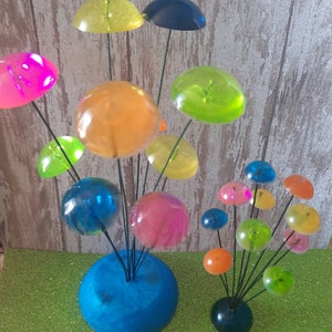 K&ouml;nnte beinhalten: Eine farbenfrohe, skurrile Skulptur aus klaren, durchsichtigen Kugeln auf Drahtstangen. Die Kugeln sind in verschiedenen Farben, darunter rosa, blau, gr&uuml;n, gelb und orange. Die Skulptur steht auf einem blauen Sockel.