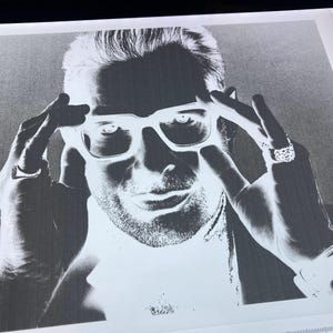 Op de afbeelding: Zwart-wit portret van een persoon met een vierkante bril, met handen die de monturen aanraken. De persoon heeft een baard en draagt een horloge en ringen. De afbeelding is een contrastrijke grijswaardenfoto.