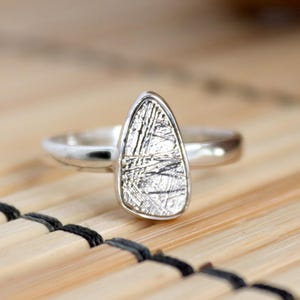 May include: A silver ring with a unique, teardrop-shaped stone. The stone has a textured, metallic appearance with a pattern of intersecting lines. The ring is set on a light-colored, natural surface.