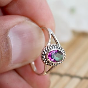 May include: A silver ring featuring an oval-cut, multi-colored gemstone. The gemstone is set within a beaded bezel. The ring's band splits into two delicate prongs that connect to the bezel. The ring is held by a hand.