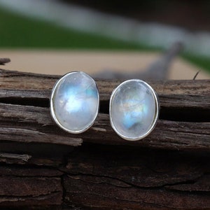 May include: A pair of silver earrings with oval-shaped moonstone gemstones. The stones have a milky white color with iridescent blue and green flashes.