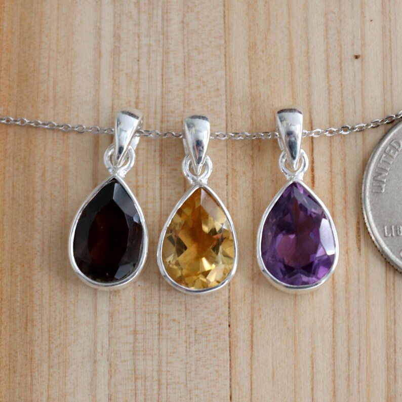 K&ouml;nnte beinhalten: Drei silberne Tropfenanh&auml;nger mit verschiedenen farbigen Edelsteinen. Die Anh&auml;nger befinden sich an einer silbernen Kette und h&auml;ngen vor einem h&ouml;lzernen Hintergrund. Die Edelsteine sind Amethyst, Citrin und Rauchquarz.