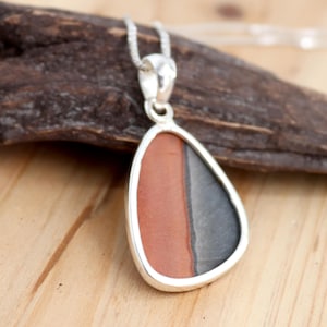 May include: A silver pendant with a teardrop-shaped stone featuring a layered pattern of red, gray, and black. The pendant is on a silver chain.