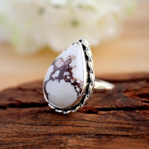 May include: A silver ring with a pear-shaped white stone, marked with brown veins. The stone is set in a decorative silver bezel. The ring is displayed on a wooden surface.