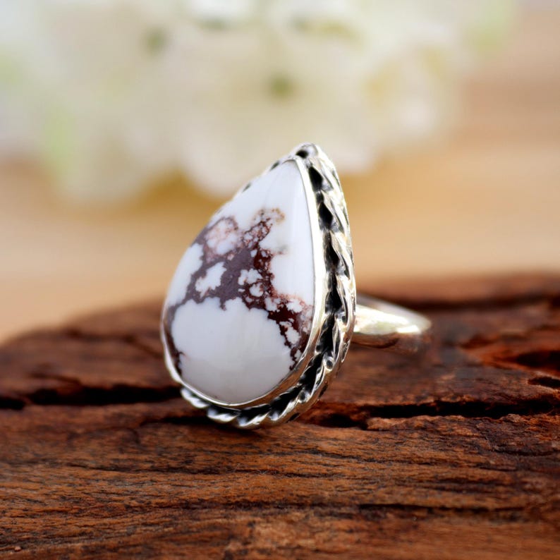 May include: A silver ring with a pear-shaped white stone, marked with brown veins. The stone is set in a decorative silver bezel. The ring is displayed on a wooden surface.