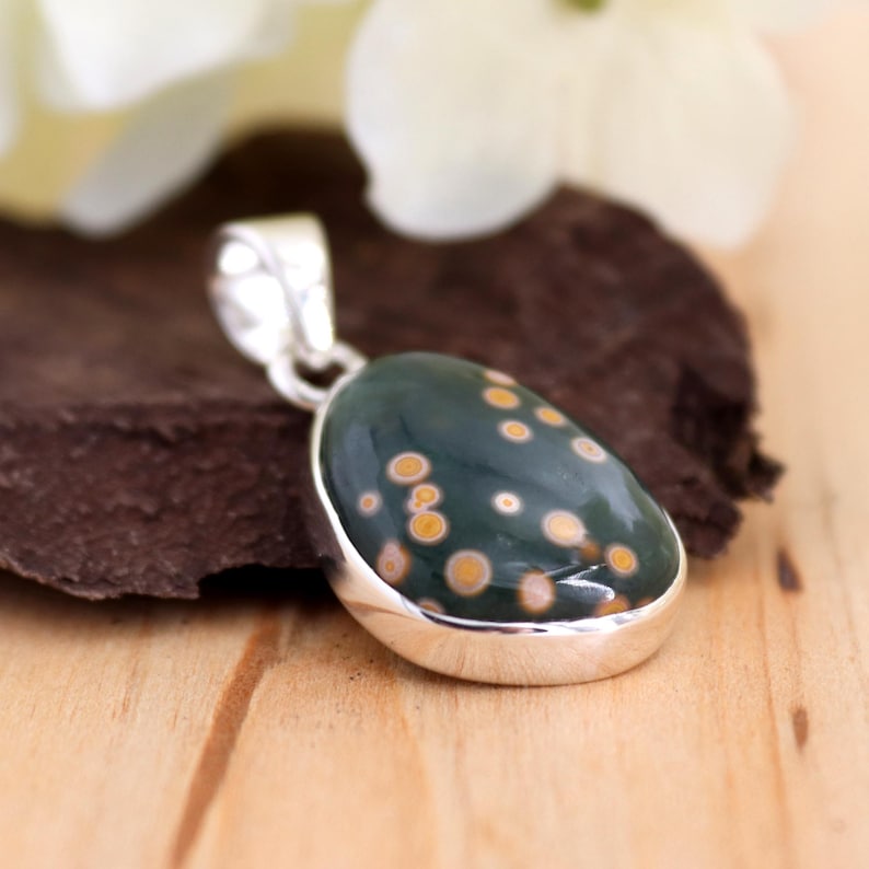 May include: A silver pendant with a green and brown ocean jasper stone. The stone has a unique pattern of small, white and yellow dots.