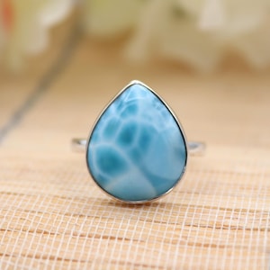 May include: A silver ring featuring a pear-shaped, light blue larimar gemstone. The stone has a unique, cloudy pattern and is set in a simple, polished silver band. The ring is displayed on a light-colored, textured surface.