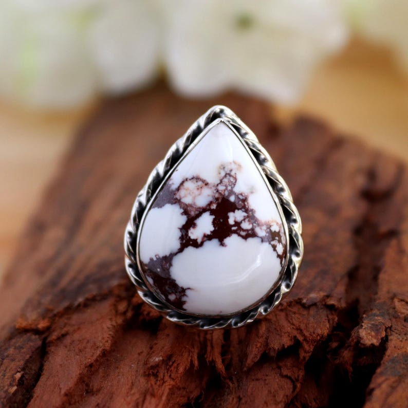 May include: A silver ring with a teardrop-shaped white stone, marbled with brown. The stone is framed by a twisted rope design. The ring is presented on a wooden surface. The ring is a piece of jewellery.