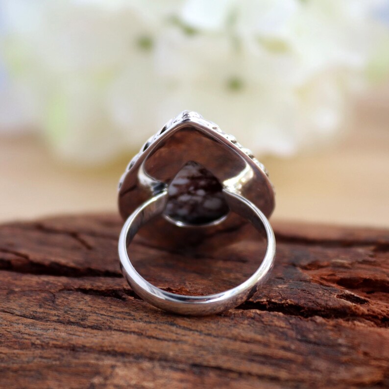 May include: A silver ring featuring a pear-shaped, speckled gemstone. The ring's band is smooth and polished, and the setting around the stone has a decorative, textured design. The ring is set on a wooden surface.