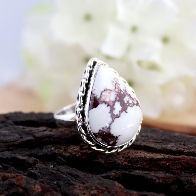 May include: A silver ring with a large, teardrop-shaped white stone, marbled with brown. The stone is set in a decorative silver bezel with a braided design. The ring is displayed on a dark, textured surface.