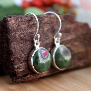 May include: A pair of silver earrings with green and pink gemstone drops. The gemstones are round and have a speckled pattern.