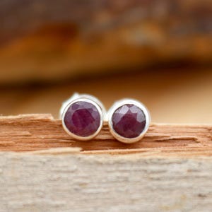 May include: A pair of stud earrings featuring faceted, deep red gemstones set in a silver bezel. The earrings are displayed on a piece of wood with a blurred, warm-toned background.