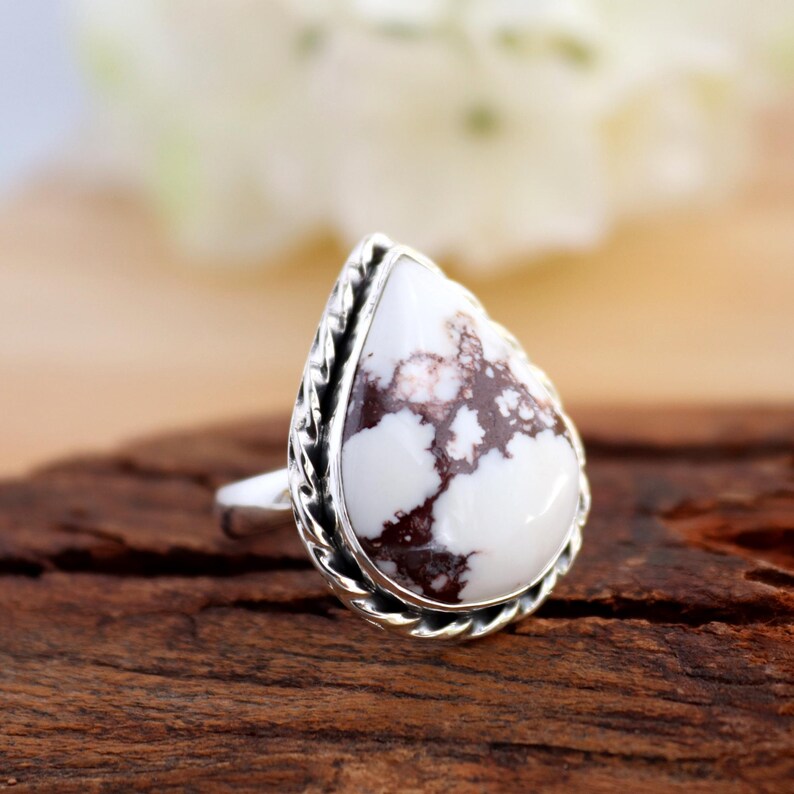 May include: A silver ring with a pear-shaped white gemstone, marbled with brown patterns. The setting has a decorative, braided edge. The ring is presented on a wooden surface, showcasing its design.