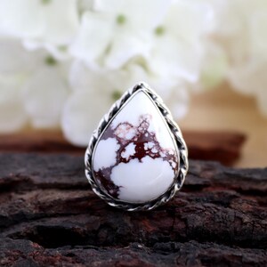 May include: A teardrop-shaped ring featuring a white stone with brown marbling, set in a silver-toned, twisted rope-like frame. The ring is displayed on a dark, textured surface, with blurred white flowers in the background.