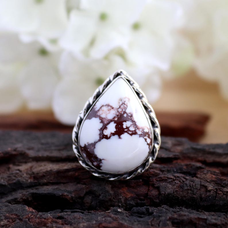 May include: A teardrop-shaped ring featuring a white stone with brown marbling, set in a silver-toned, twisted rope-like frame. The ring is displayed on a dark, textured surface, with blurred white flowers in the background.