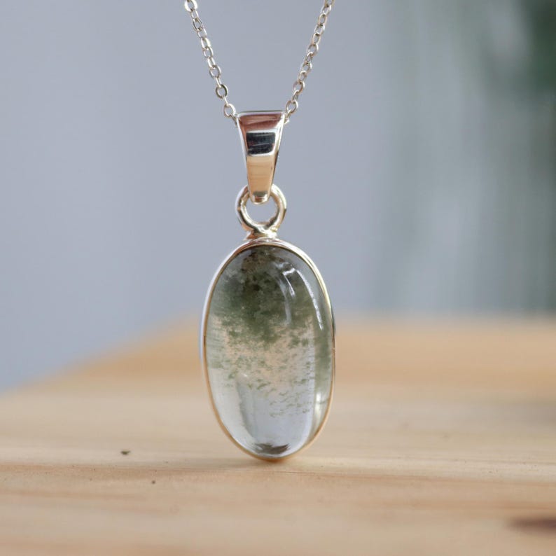 May include: A silver chain necklace with a green and white oval pendant. The pendant is made of a clear stone with green inclusions.
