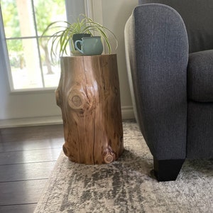 May include: A wooden stump side table with a natural wood finish. The table has a rough, textured surface and is decorated with two small ceramic mugs. The table is sitting on a light grey rug with a patterned design.