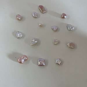 May include: A collection of 13 unique, irregular shaped pearls in various shades of white, pink, and beige. The pearls are arranged on a white surface.