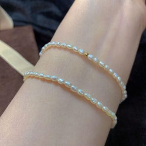 May include: Two delicate pearl bracelets, each featuring small, oval-shaped pearls strung together. A small gold bead accents one bracelet. The bracelets are worn on a wrist, showcasing the jewelry's elegant design and subtle luster.