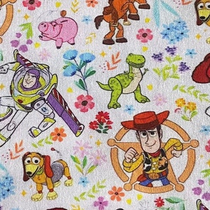 May include: White fabric featuring a repeating pattern of Toy Story characters such as Woody, Buzz Lightyear, and Jessie. The design is interspersed with colourful flowers and other decorative elements.
