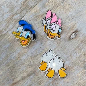 May include: Four cartoon character iron-on patches. Two are of Donald Duck with a blue hat and a yellow beak. Two are of Daisy Duck with a pink bow and a yellow beak. The patches are glittery.