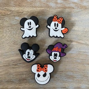May include: Five Halloween-themed shoe charms featuring cartoon characters. The charms include a ghost, a witch, and a character with a pumpkin. The charms are black, white, orange, and purple, and are arranged on a wooden surface.