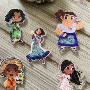 May include: Five colorful character acrylic charms from the movie Encanto. The charms feature detailed illustrations of the characters, with glitter accents. The characters are in various poses, with vibrant clothing and accessories.