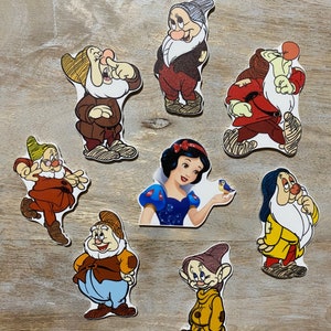 May include: Seven colorful cartoon characters from the fairy tale Snow White and the Seven Dwarfs. The characters are all standing on a wooden surface. The characters are all wearing different colored clothing. The characters are all smiling.
