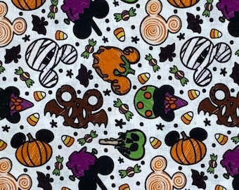 May include: White fabric with a repeating pattern of Halloween-themed Mickey Mouse shapes, pumpkins, bats, candy corn, and skulls. The design features orange, purple, green, and brown elements.