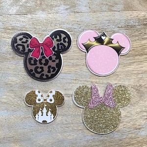Peut inclure: Quatre découpes décoratives en forme de tête de souris. L'une présente un motif léopard avec un nœud rouge. Une autre a un fond rose avec une couronne dorée. Les deux autres ont des paillettes dorées avec un château blanc et un nœud rose.
