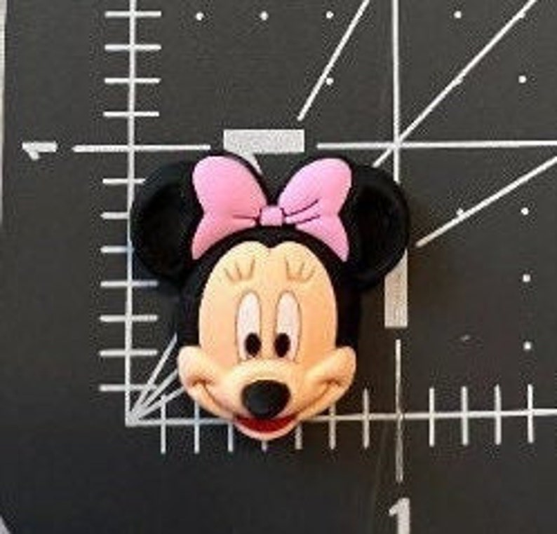 May include: A cartoon character head of Minnie Mouse with black ears, a pink bow, and a smiling face.