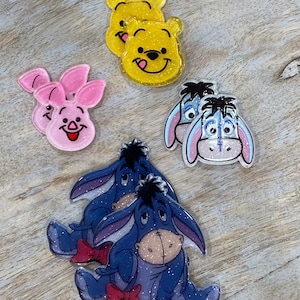 May include: A set of six colorful character-themed resin charms. The charms feature Winnie the Pooh, Piglet, and Eeyore from the Disney cartoon. The charms are made of clear resin with glitter embedded in the resin.