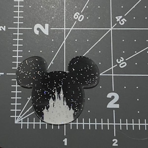 Puede incluir: Orejas de Mickey Mouse negras y blancas con purpurina y una silueta blanca del castillo de Cenicienta en el centro.