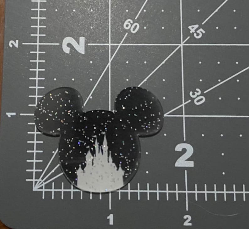 Puede incluir: Orejas de Mickey Mouse negras y blancas con purpurina y una silueta blanca del castillo de Cenicienta en el centro.