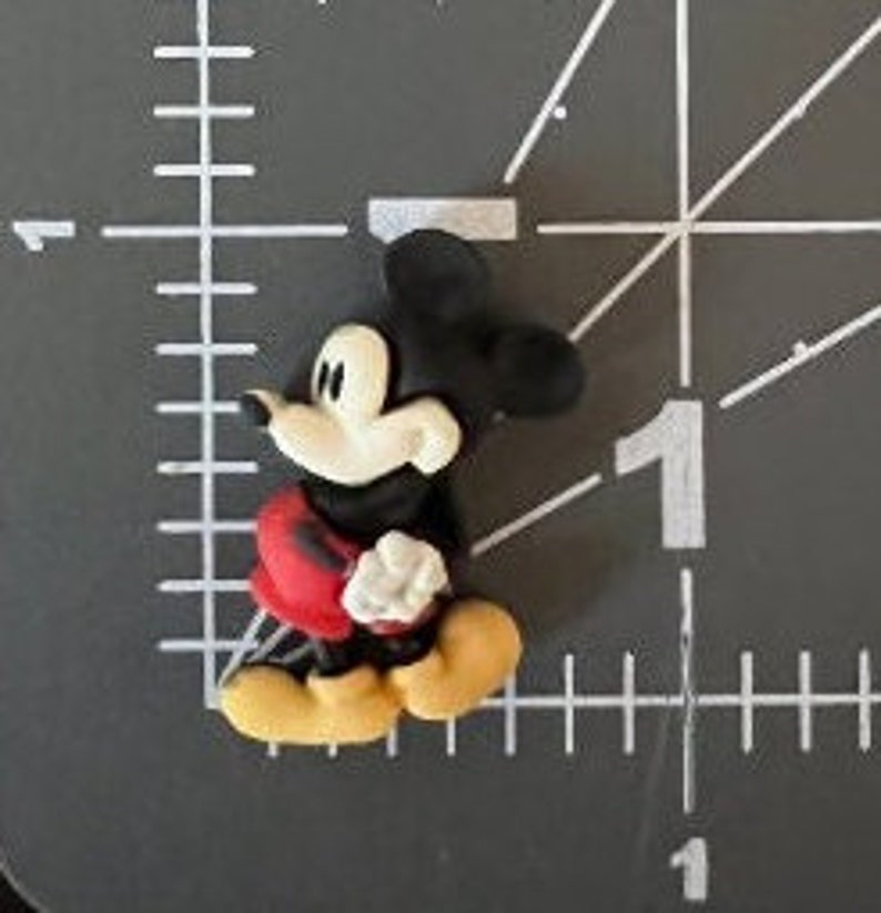 Puede incluir: Una peque&ntilde;a figura tridimensional de Mickey Mouse. La figura tiene orejas negras y cabeza negra, cara blanca, pantalones cortos rojos, zapatos amarillos y guantes blancos. La figura est&aacute; sobre una superficie gris con una regla.