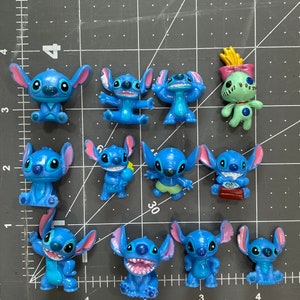 May include: A collection of 11 blue Stitch figurines, including a green Angel figurine. The Stitch figurines are in various poses, some holding objects like a flower and a book. The figurines are made of plastic and have a glossy finish.