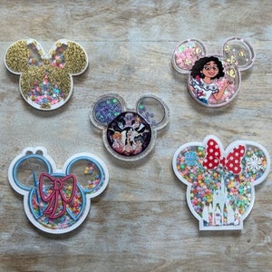 May include: Five different mouse-shaped resin ornaments with glitter and various images inside. The ornaments are white with black outlines and feature a red bow, a castle, a group of Disney characters, a butterfly, and a woman with a flower crown.