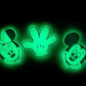Puede incluir: Tres diseños de personajes de dibujos animados de color verde brillante sobre un fondo negro. Los diseños incluyen Mickey Mouse, Minnie Mouse y una mano enguantada. Los personajes y la mano están delineados con un brillo verde brillante.